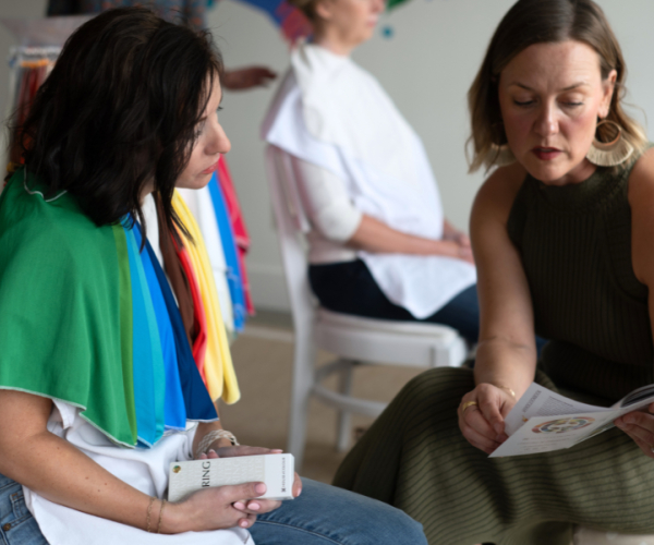 Two individuals seated indoors during a colour analysis session, with one person holding a fan of colour swatches draped across their shoulders and another holding a booklet. Additional colour drapes are visible in the background.